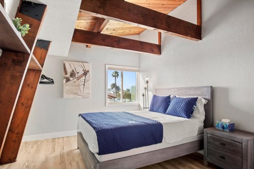 Cozy bedroom with a wooden ceiling, large window, and a bed with blue bedding, featuring modern decor and natural light.
