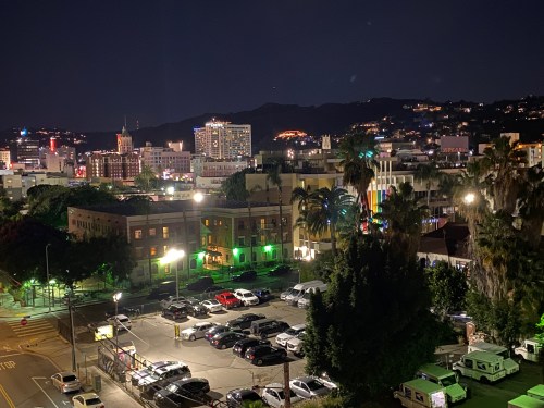 Nighttime cityscape featuring illuminated buildings, palm trees, and a parking lot in the foreground.