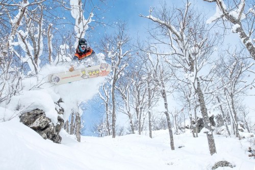 A snowboarder jumps off a snowy slope surrounded by bare trees under a clear blue sky.