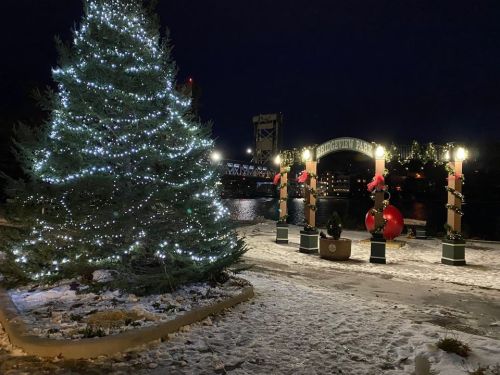 A decorated Christmas tree and festive archway in a snowy park at night, with a bridge and water in the background.