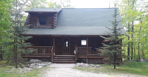 A rustic log cabin surrounded by trees, featuring a front porch and a gravel pathway leading to the entrance.