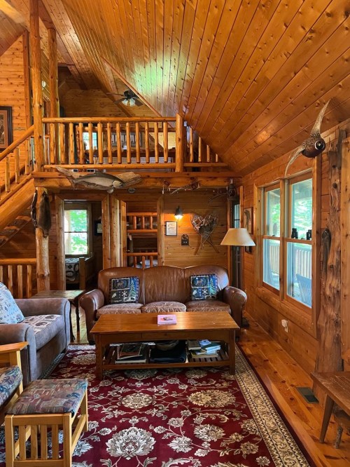 Cozy wooden cabin interior with a leather sofa, coffee table, and rustic decor, featuring a loft and large windows.