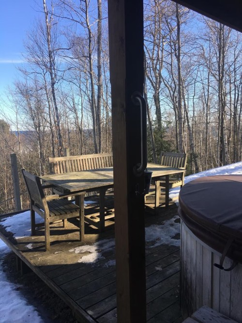 A wooden deck with a table and chairs, surrounded by trees and snow, under a clear blue sky.