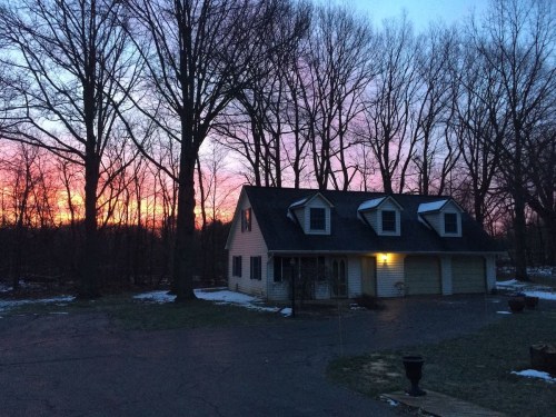 A house with a garage surrounded by trees at sunset, with vibrant pink and orange skies and some snow on the ground.
