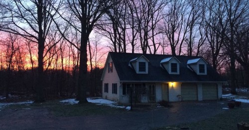 A cozy house with a sloped roof sits in a wooded area, illuminated by a vibrant sunset sky. Snow is visible on the ground.