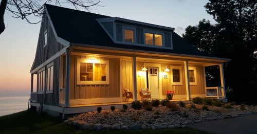 A cozy, illuminated house at dusk, featuring a porch, large windows, and a scenic view of the water.