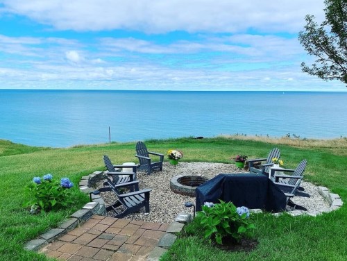 A serene lakeside view featuring a fire pit surrounded by chairs and blooming flowers under a blue sky.