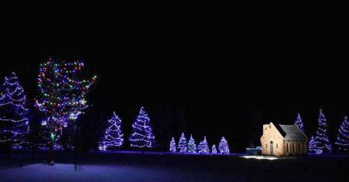 A snowy landscape at night featuring a small house and colorful, illuminated trees.