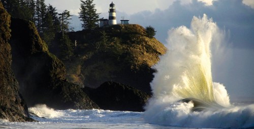 A lighthouse on a rocky shore with waves crashing against the rocks under a cloudy sky.