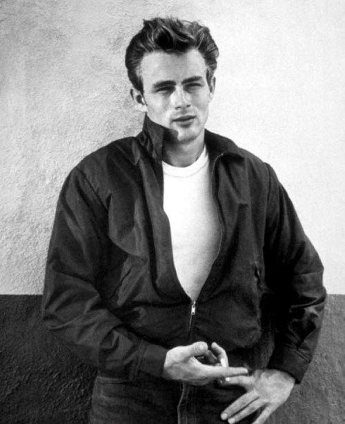 A young man in a black jacket and white shirt stands against a wall, looking confidently at the camera.