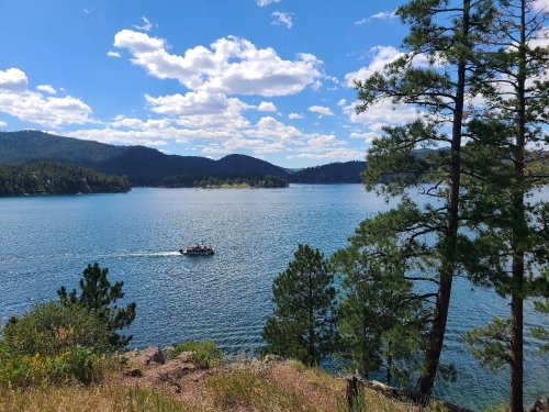 A scenic view of a lake surrounded by mountains and trees, with a boat gliding across the water under a blue sky.