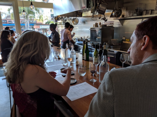A couple sits at a bar in a restaurant, reviewing menus while a chef prepares food in the background.