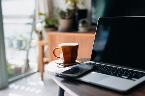 A laptop and smartphone on a table, with a brown mug of tea or coffee beside them in a cozy, sunlit room.