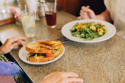 A close-up of a sandwich on a plate next to a salad, with hands visible at a table in a diner setting.