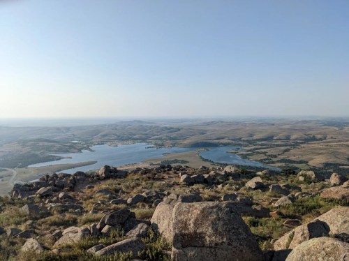 A panoramic view of rolling hills and a lake from a rocky vantage point under a clear blue sky.