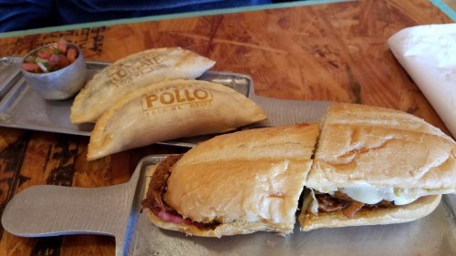 A cut sandwich filled with meat and cheese, served with two empanadas and a small bowl of salsa on a wooden table.