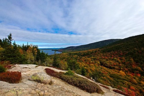 A scenic view of a colorful autumn landscape with trees, mountains, and a lake under a partly cloudy sky.