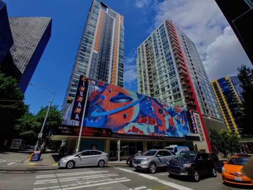 A vibrant cinema building with colorful murals, surrounded by tall modern skyscrapers and parked cars on a sunny day.