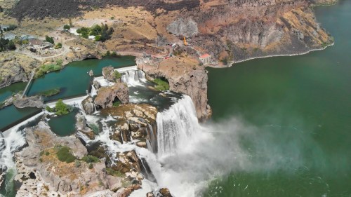 Aerial view of a large waterfall cascading into a green river, surrounded by rocky terrain and lush vegetation.