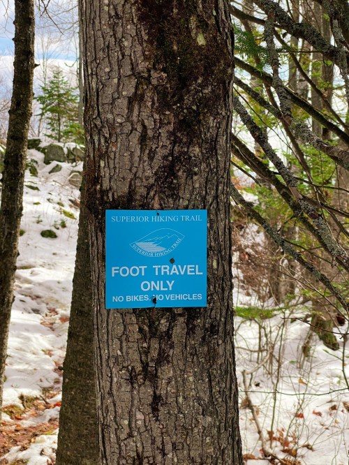 A blue sign on a tree reads "Foot Travel Only" with a note about no bikes or vehicles allowed on the Superior Hiking Trail.