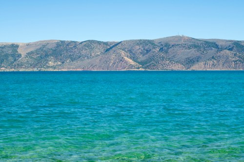 A clear blue lake with gentle waves, surrounded by green hills under a bright blue sky.