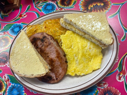 A plate with two slices of buttered bread, ham, scrambled eggs, and hash browns on a colorful floral tablecloth.