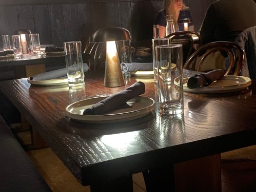 A dimly lit restaurant table set with plates, glasses, and rolled napkins, with a warm lamp illuminating the scene.