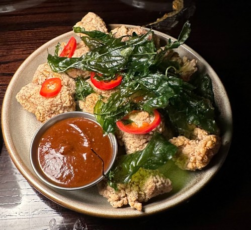 A plate of crispy fried pieces with green leaves and red chili slices, accompanied by a small bowl of sauce.