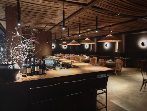 A modern restaurant interior featuring a wooden bar, wine bottles, and stylish pendant lighting over dining tables.