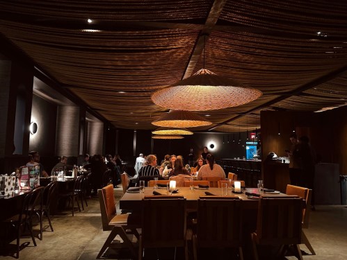 A dimly lit restaurant interior with woven ceiling decor, featuring tables and patrons enjoying their meals.
