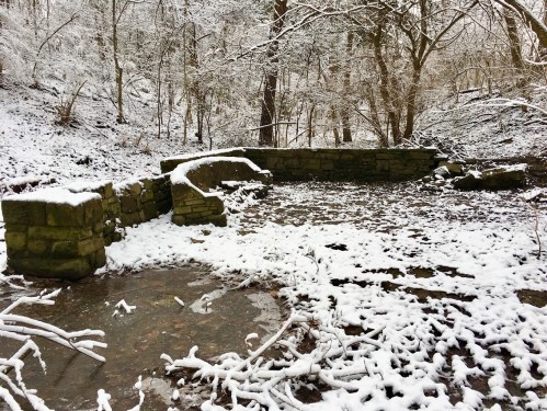 A snowy landscape featuring a stone structure surrounded by trees and a blanket of white snow on the ground.