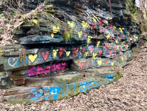 Colorful heart graffiti in red, yellow, and blue on a rocky wall, surrounded by dry leaves and greenery.