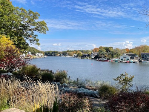 A serene river view with boats, lush greenery, and colorful autumn trees under a blue sky with fluffy clouds.