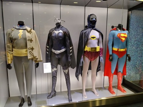 Four superhero costumes displayed on mannequins: a gray suit, a black bodysuit, a Batman outfit, and a Superman costume.