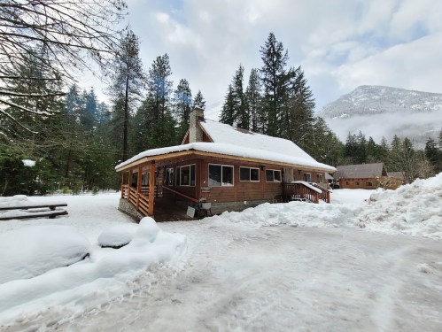 A cozy wooden cabin surrounded by snow-covered trees and mountains under a cloudy sky.