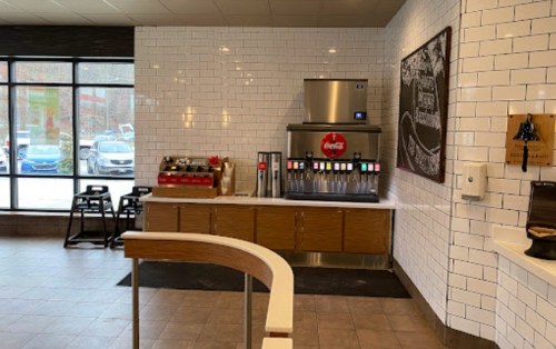A beverage station with a Coca-Cola dispenser, surrounded by white tiles and wooden accents in a bright, modern space.