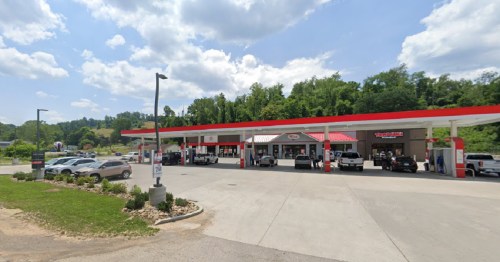 A gas station with several vehicles at the pumps, surrounded by greenery and under a partly cloudy sky.