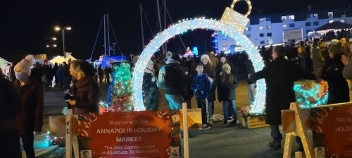 A festive holiday market in Annapolis, with a large illuminated arch and crowds enjoying the decorations and activities.