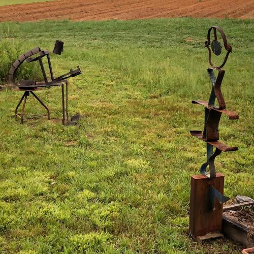 Two abstract metal sculptures in a grassy field, with a plowed field in the background.