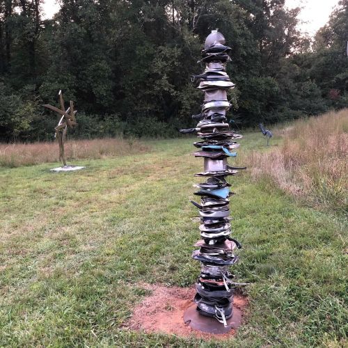 A tall sculpture made of stacked metal plates stands in a grassy field, surrounded by trees and another sculpture in the background.