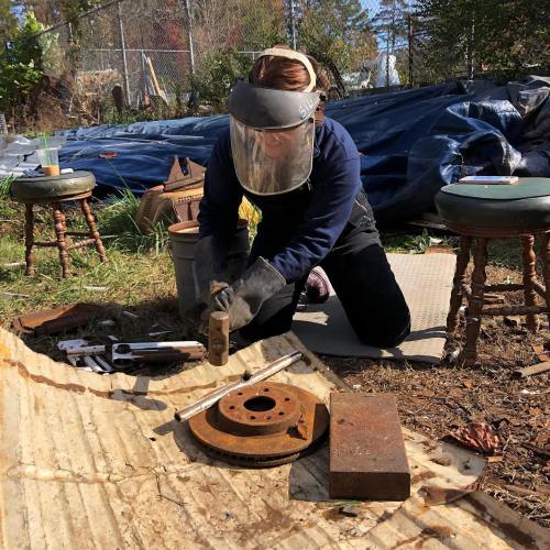 A person wearing a protective face shield works on a metal object outdoors, surrounded by tools and materials.