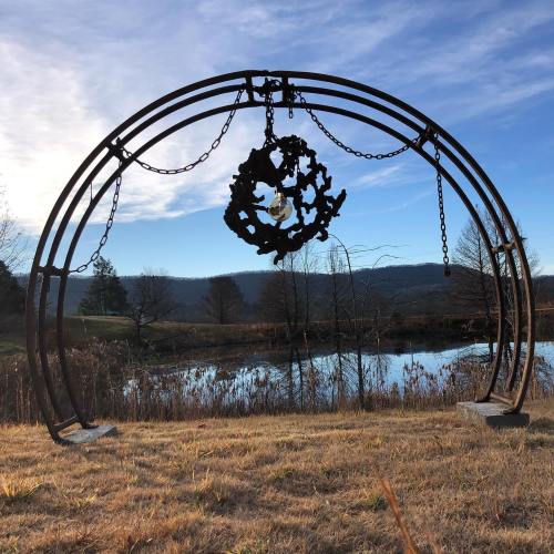 A metal arch with a circular design stands near a calm pond, surrounded by trees and a clear sky.