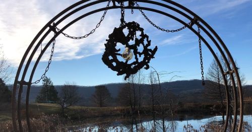 A metal sculpture hangs in a circular frame, overlooking a serene pond and distant hills under a clear blue sky.