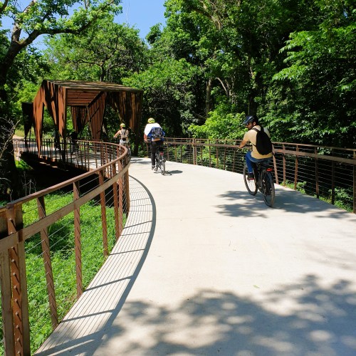 Two cyclists ride along a curved path surrounded by lush greenery, with a wooden bridge in the background.