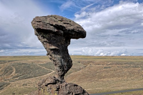 A tall, mushroom-shaped rock formation stands against a backdrop of rolling hills and a cloudy sky.