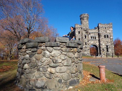 A person stands on a stone structure in front of a castle-like building, surrounded by autumn trees and a clear blue sky.