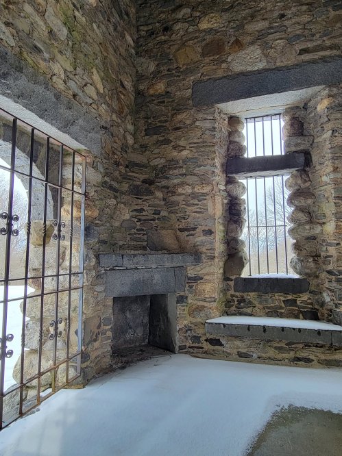 Interior of a stone structure with barred windows, a fireplace, and snow on the floor, creating a cold, abandoned atmosphere.