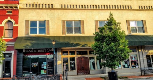 A street view of a brick building with large windows and a green awning, featuring a nearby café and outdoor seating.
