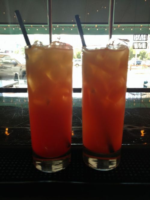 Two tall glasses filled with a colorful iced drink, featuring ice cubes and black straws, on a bar counter.