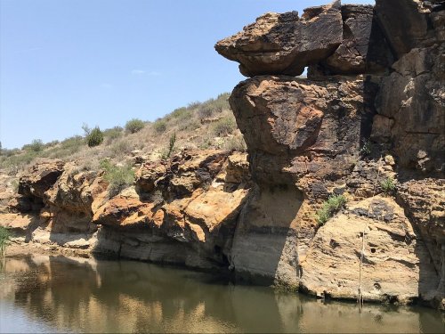 A rocky cliff with unique formations reflected in calm water, surrounded by sparse vegetation under a clear blue sky.